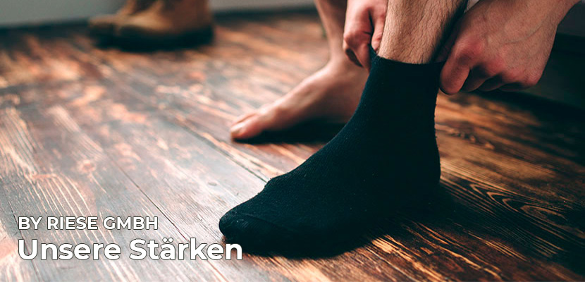 by Riese GmbH & Co. KG Titelbild RS Harmony - Titelbild der Startseite von by Riese GmbH & Co. KG - Socken und Underwear. Ein Mann zieht sich einen schwarzen Socken über den Fuß. Im Hintergrund stehen im verschwommenen seine Schuhe.